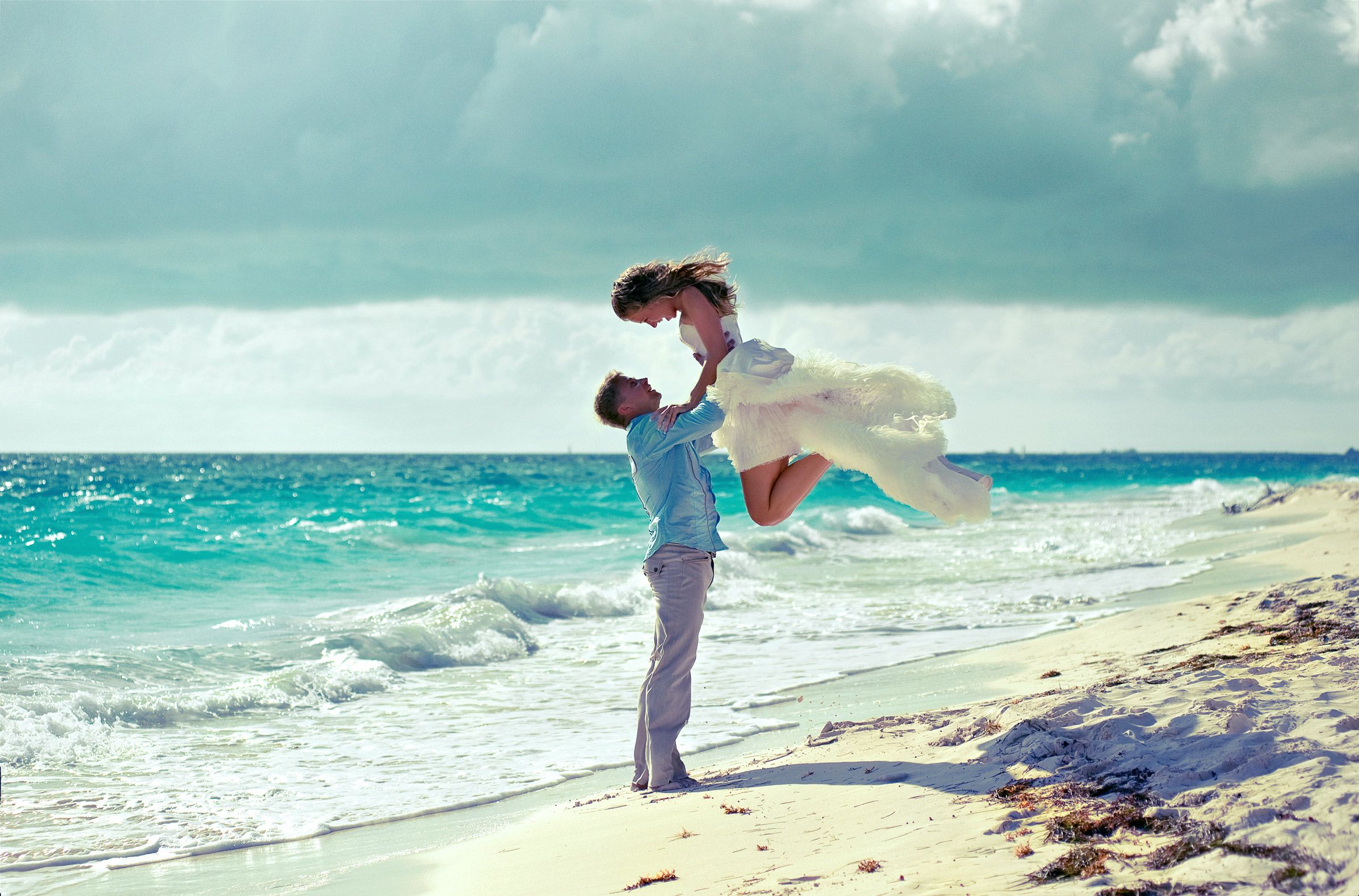 Groom lifts bride joyfully on a sandy beach with turquoise waves behind them.