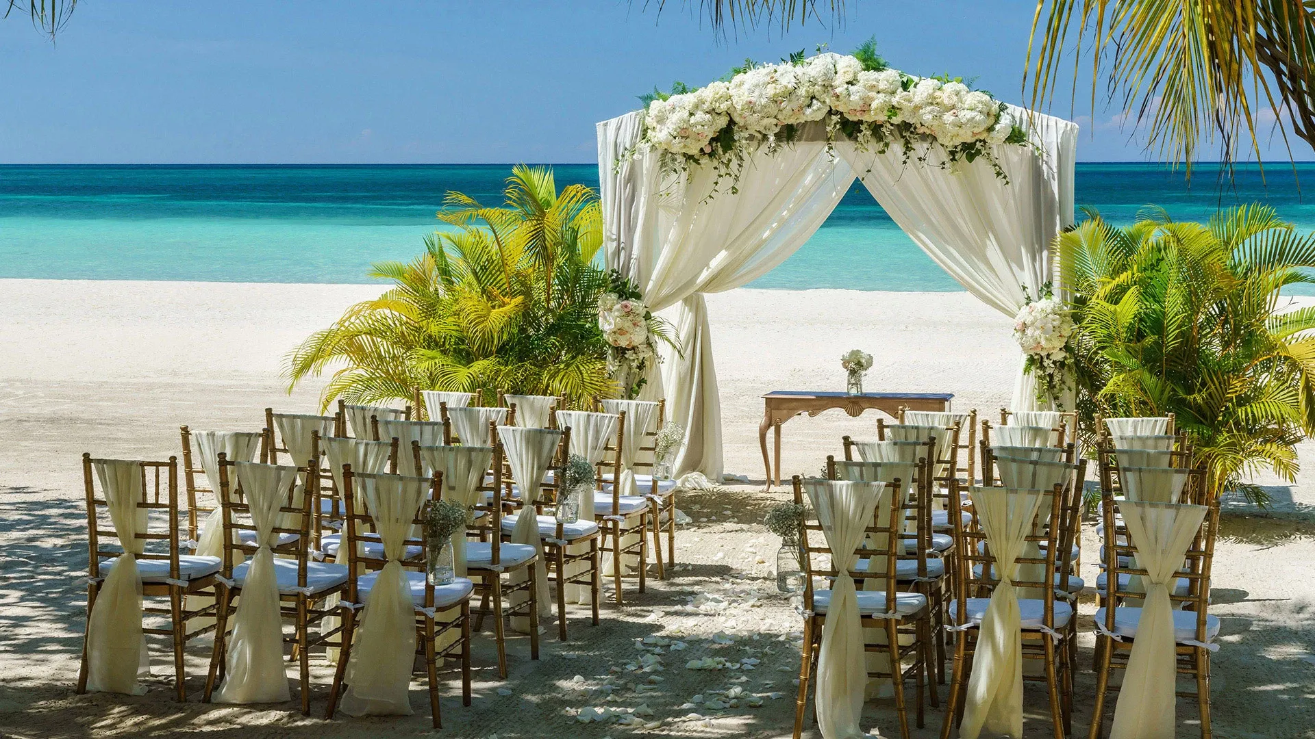 Elegant beachfront wedding ceremony setup with white floral arch and wooden chairs.