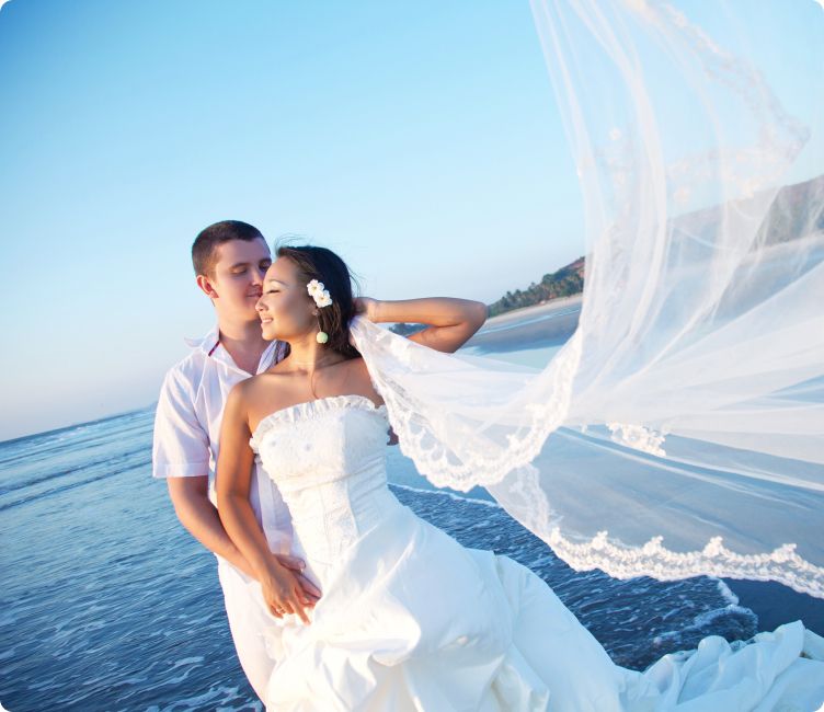 Bride and groom stand on the beach with veil flowing dramatically in the breeze.