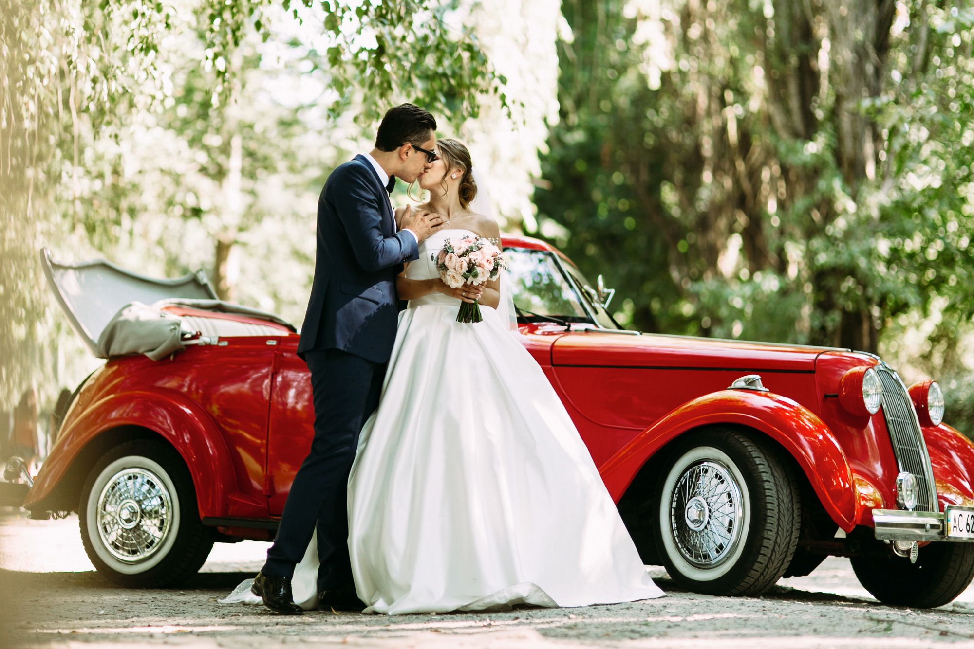 Bride and groom kissing in front of a red vintage convertible in a wooded area.