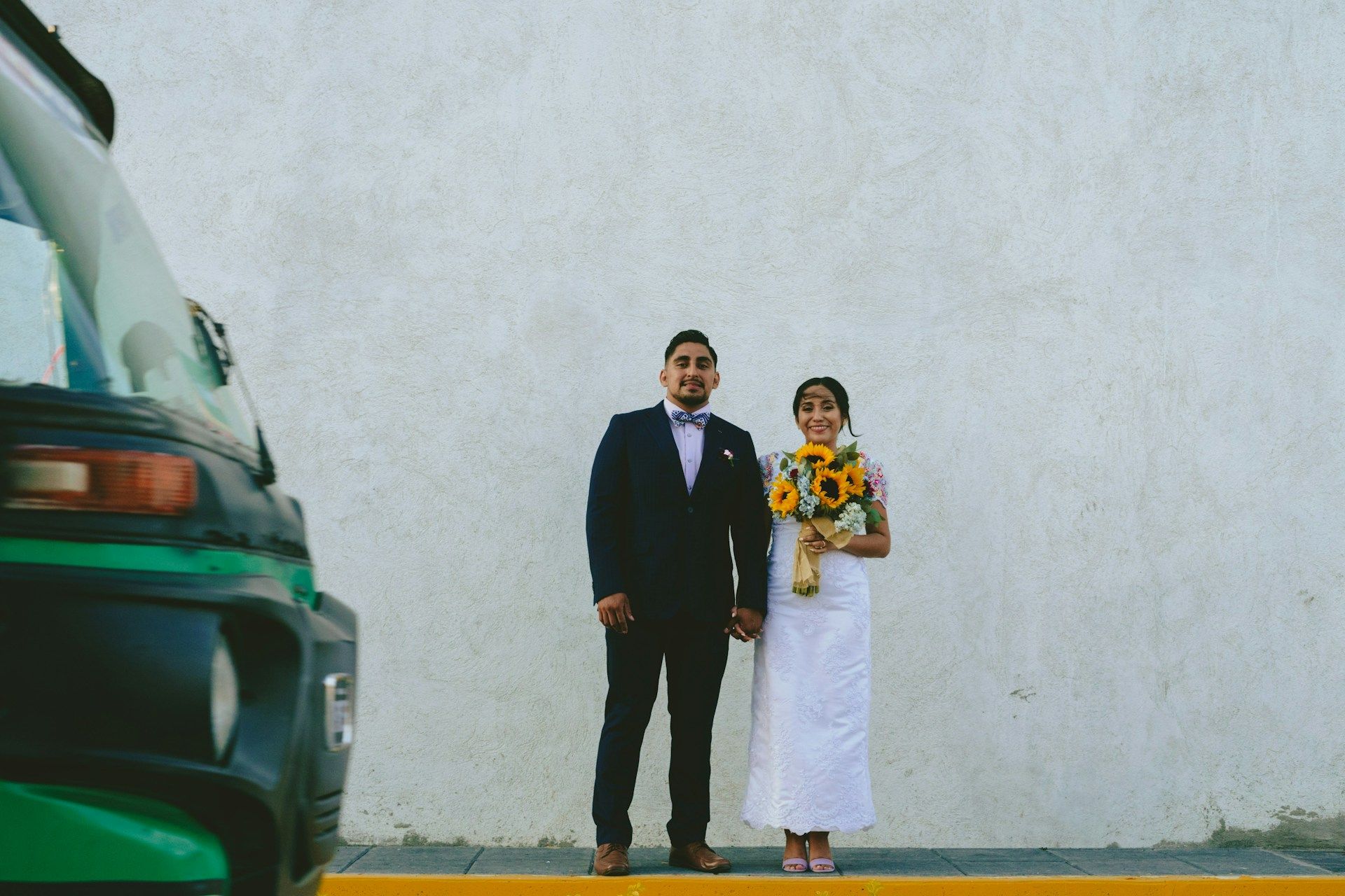 Bride and groom standing together against a white wall, holding hands.