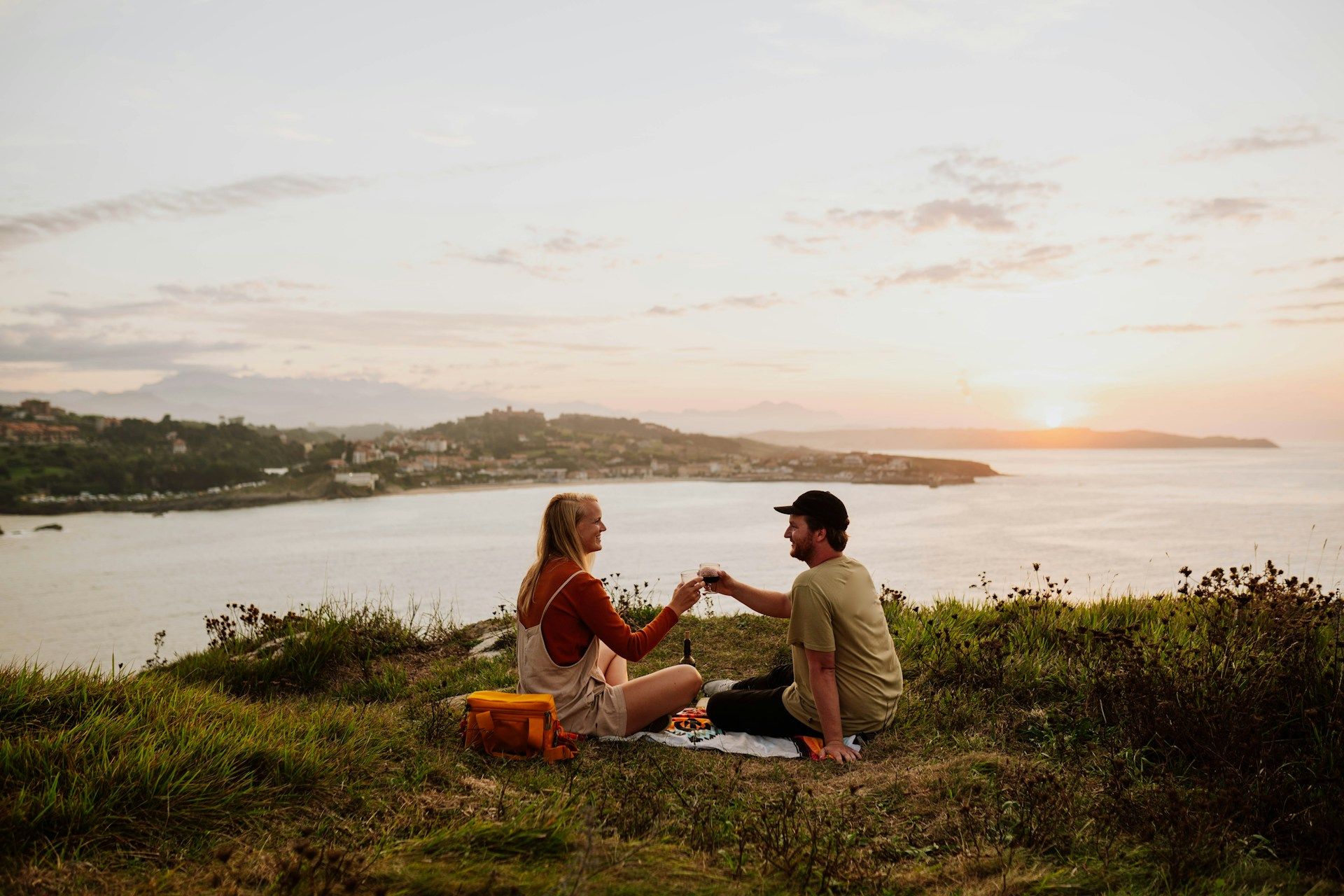 couple cheering glasses and enjoying a picnic on grassy cliff overlooking the water