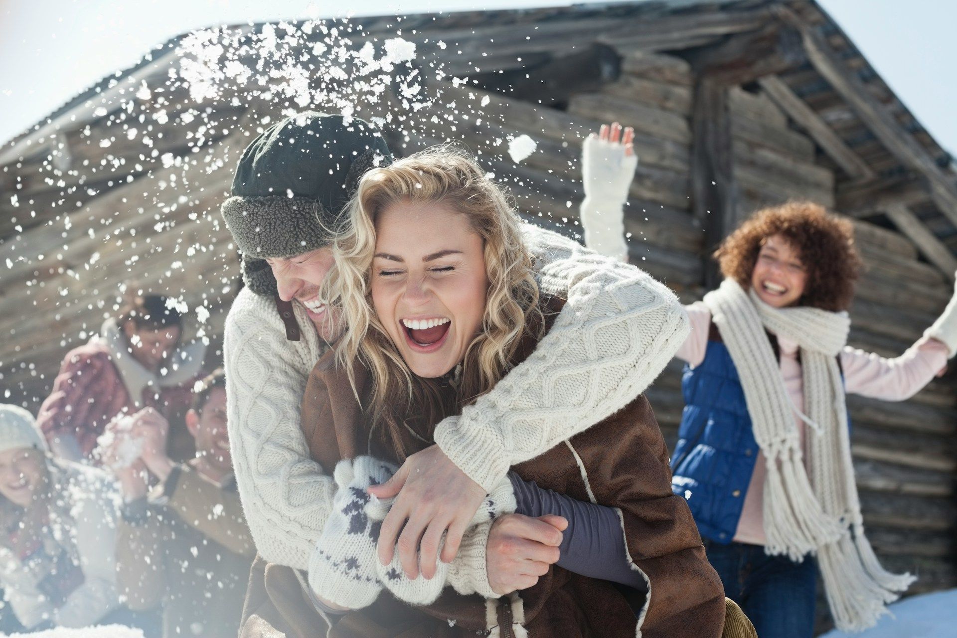 Friends playing in the snow and laughing outside a wooden cabin.