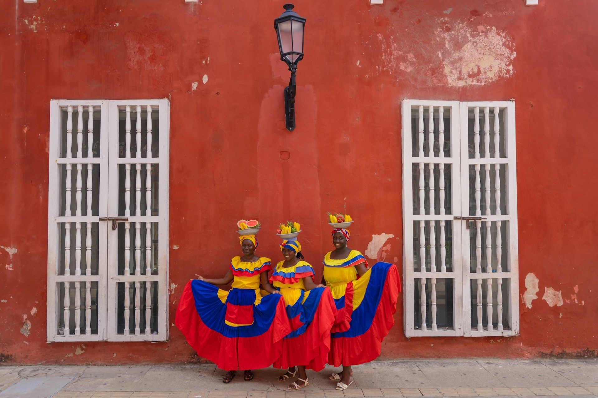 Three women in colorful traditional dresses pose against a red wall, holding fruit baskets on their heads.