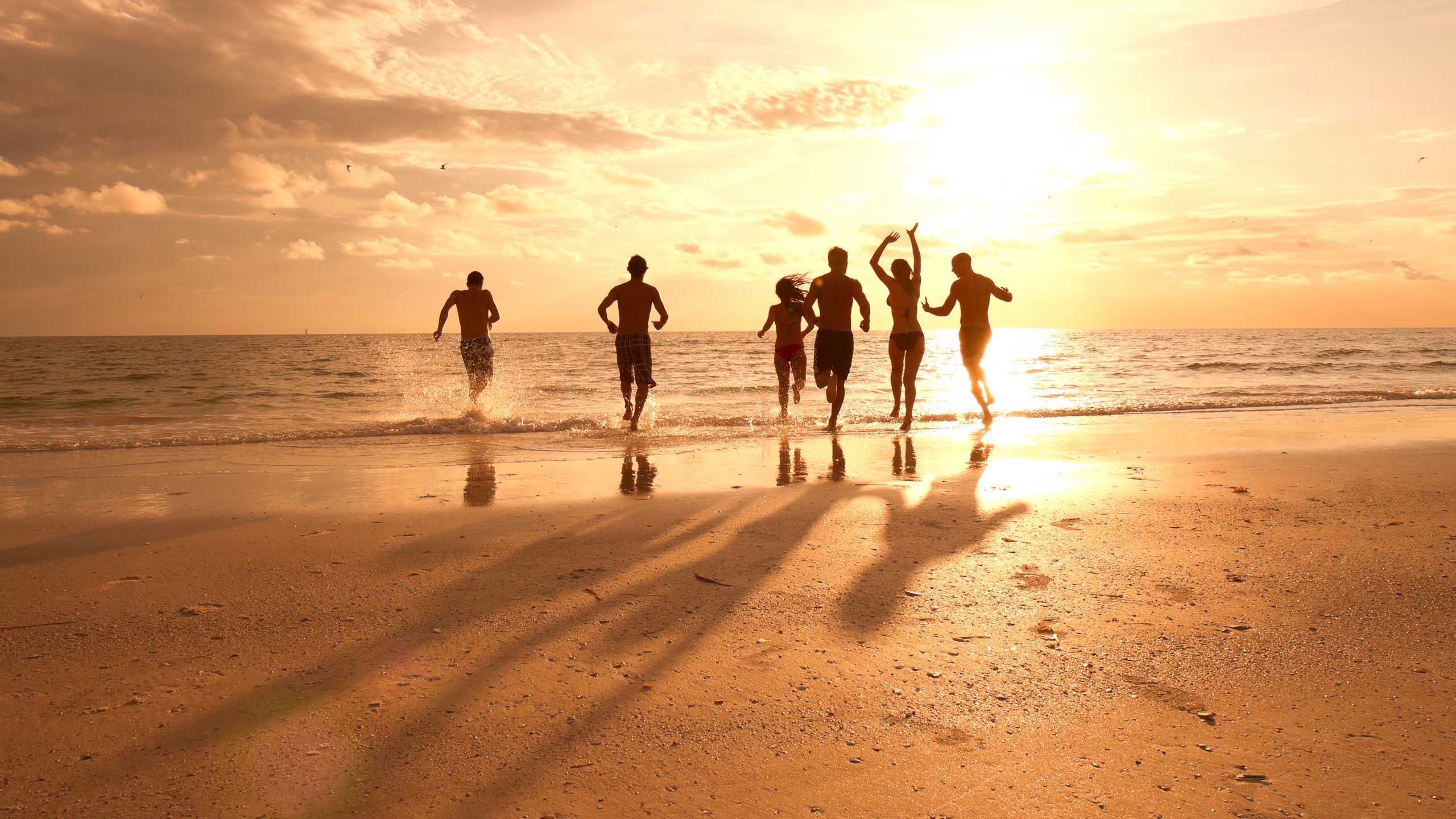 Friends running into the ocean together at sunset, casting long shadows on the beach