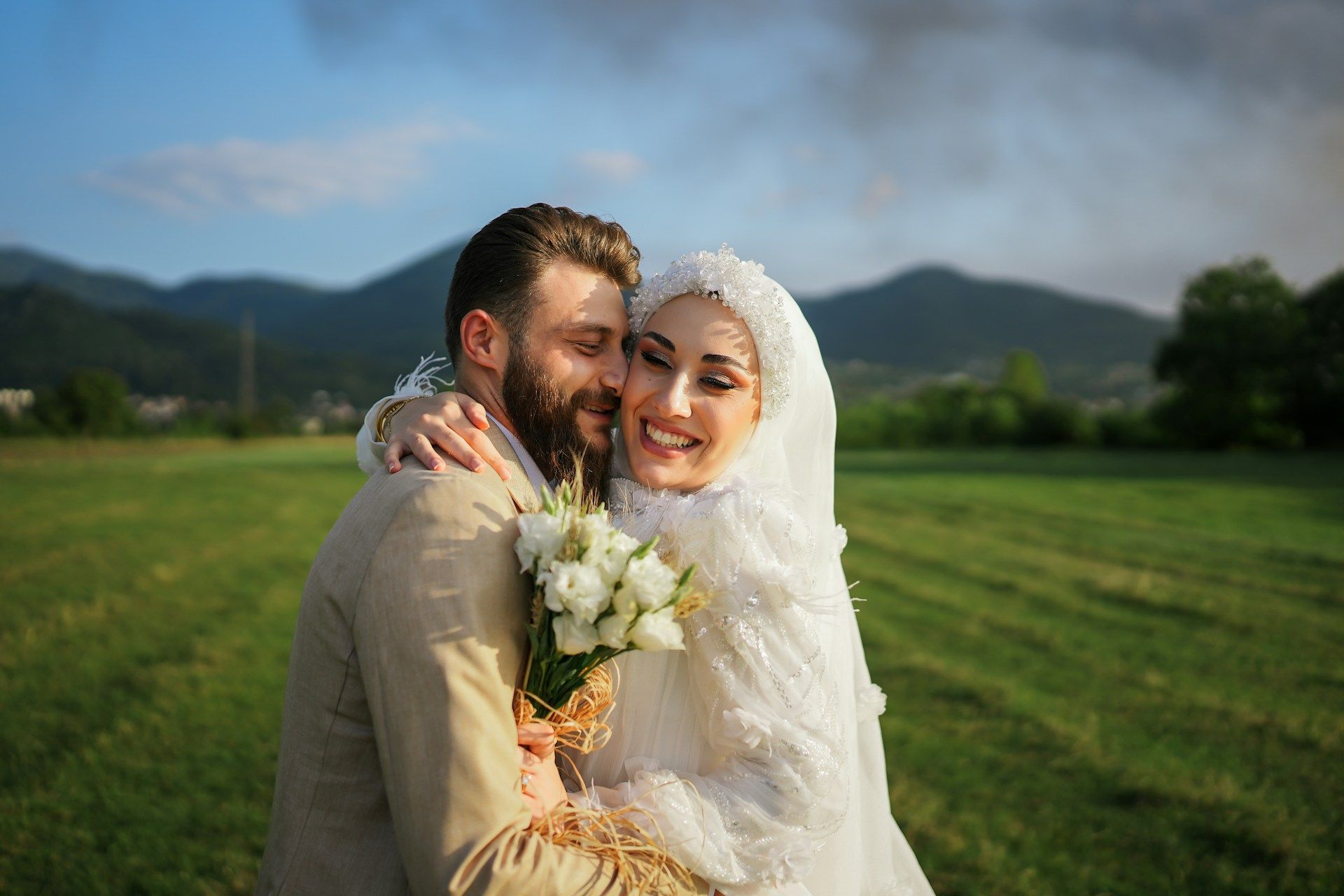 Smiling bride and groom hugging in a green field with mountains in the background.